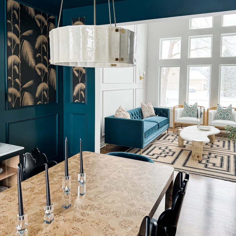 An image of the reimagined spaces designed by the Firelight Style House interior design team. The image shows a view from a dining room into a living room that has many large windows, all showing a snowy landscape in the background. The foreground is a sleek modern table, crystal candle holders, and a new light fixture that hangs from the ceiling. The dining room is in a cool dark green-navy color, and some panels have wallpaper that is black with a cream silhouette of plants. The team also reimaged the living room and features a bright green couch that pairs well with the color of the walls in the dining room, two chairs, a contemporary table, and a cream-colored rug with a black design on it.