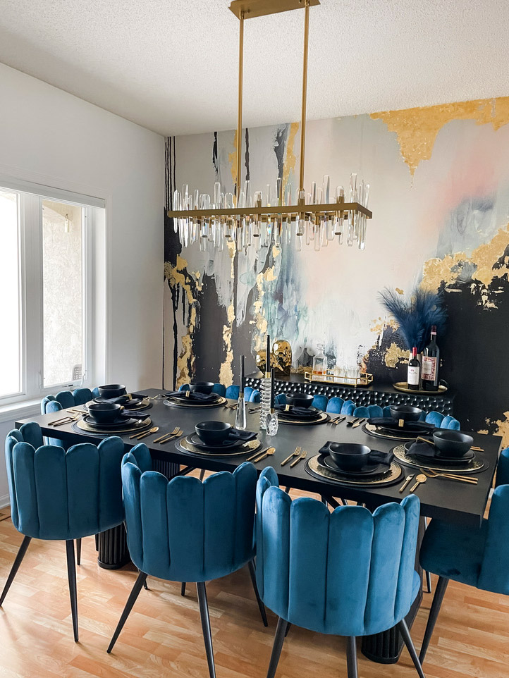 This dining room in the Minneapolis suburbs showcases a striking modern design with teal velvet scalloped dining chairs around a black rectangular table. A gold and glass linear chandelier hangs overhead, while the abstract mural with navy, gold, and blush tones brings vibrant movement to the space.