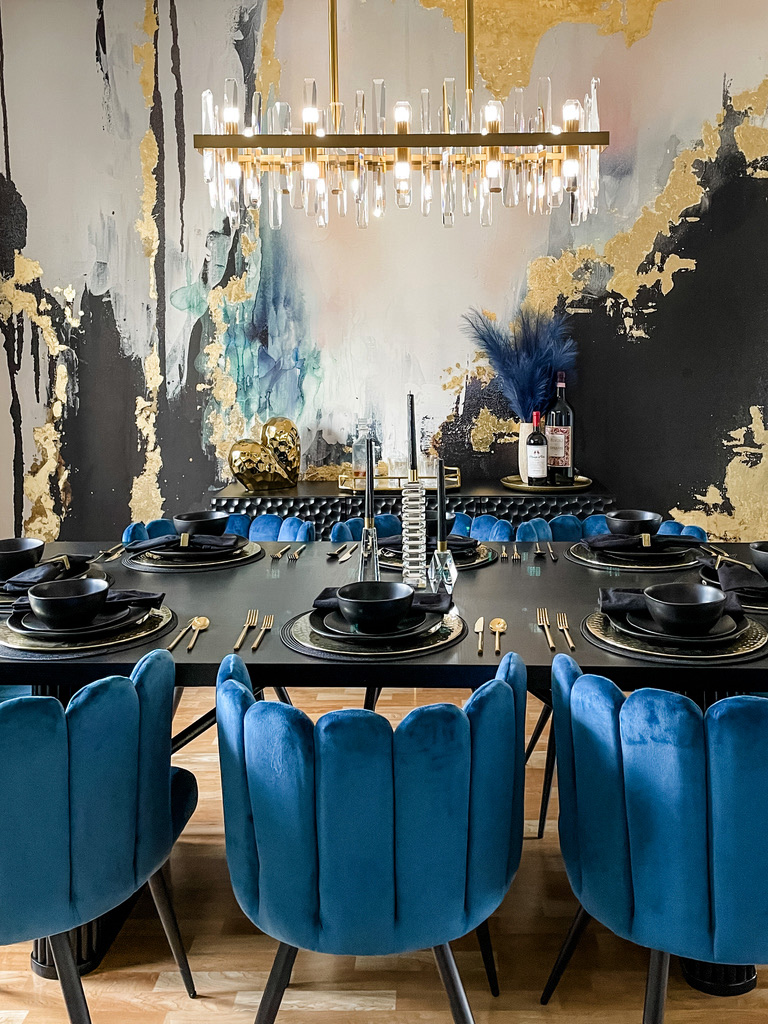 A close-up view of the Minneapolis-St. Paul dining room highlights the vibrant blue velvet chairs and black tabletop set with matte black dishware and gold flatware. Behind the table, a stunning wall mural in metallic gold and navy adds a custom artistic flair. This expressive dining room reflects the design team's boutique vision—merging custom art-forward pieces with elevated entertaining spaces for clients across the Twin Cities.
