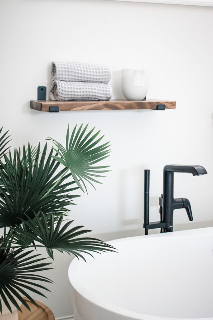 Close-up of a matte black tub filler paired with a white soaking tub, topped with a wooden bath tray and neutral-toned accessories. A palm leaf plant and minimalist floating shelf bring organic elements to this Minneapolis-St. Paul bathroom design. The interior design team's attention to calming textures and natural styling reflects their boutique sensibility and custom approach.