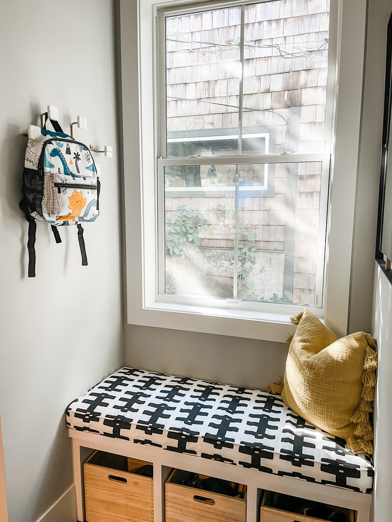 A cheerful mudroom nook with a window view of cedar shake siding from an adjacent home in the Minneapolis suburbs. A built-in bench features a black-and-white patterned cushion, accented with a mustard-yellow throw pillow. Beneath the bench are woven storage bins, and a children’s backpack hangs from wall-mounted white hooks—practical and stylish family-friendly design.