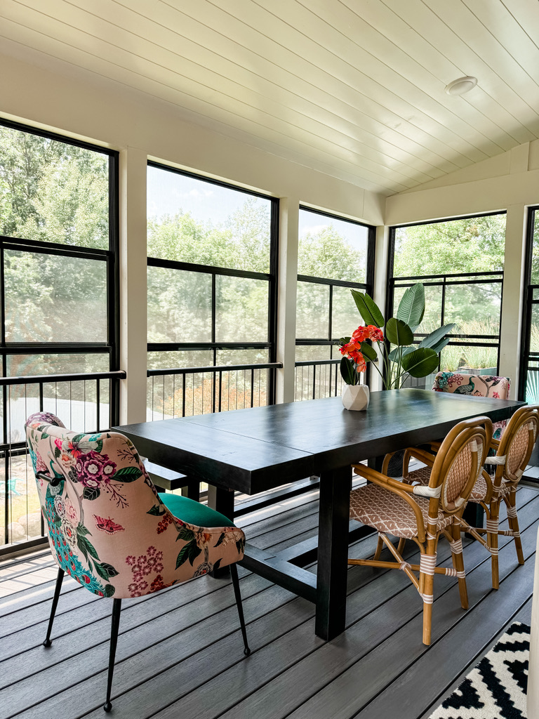 A sunroom dining space in the Minneapolis suburbs with black-framed floor-to-ceiling windows that let in ample natural light. The room features a long black wood dining table surrounded by woven rattan chairs and a pair of bold floral accent chairs with peacock and blossom prints. A white sculptural vase with vivid red flowers and a tall leafy plant add color and height, bringing a lively, garden-inspired elegance to the space.