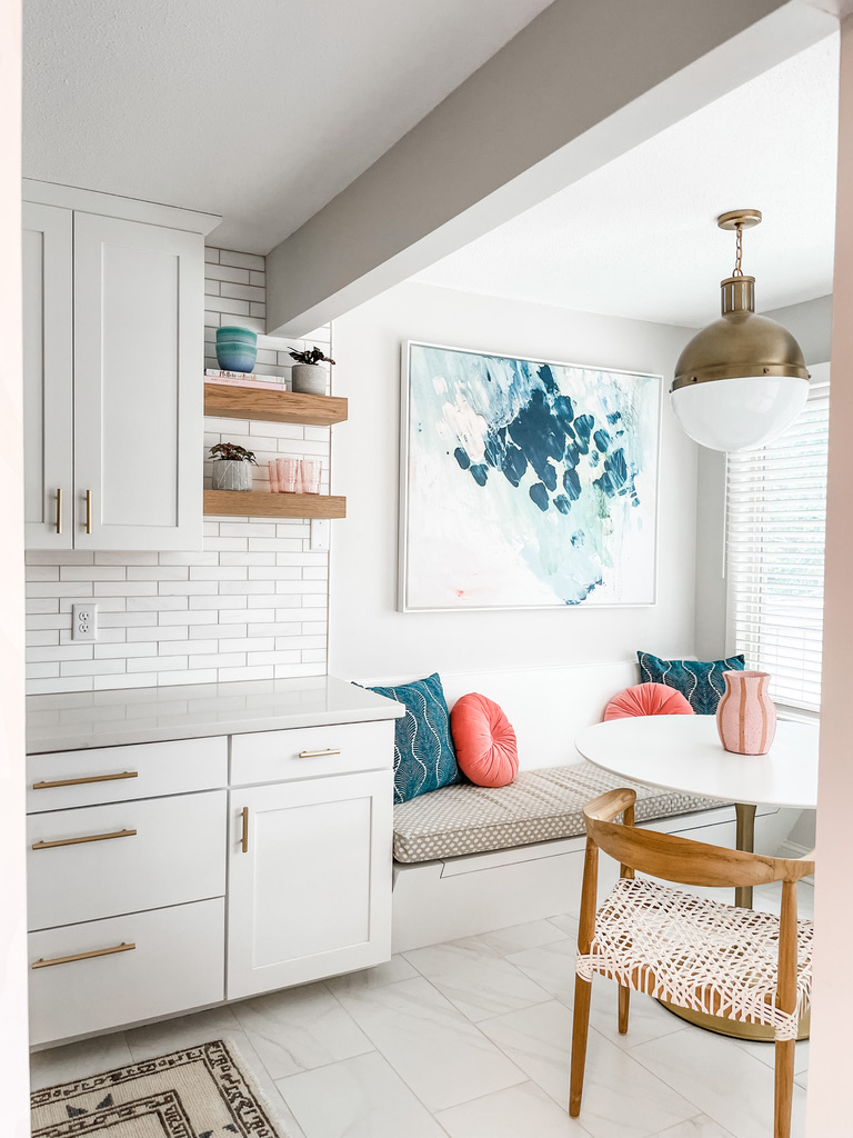 A sunny kitchen nook showcases crisp white cabinetry with gold pulls, a built-in bench upholstered in neutral patterned fabric, and accent pillows in teal and coral hues. A round white table sits beside a woven blush and white chair. Overhead, a brass and globe pendant light adds mid-century charm, and a watercolor-style abstract painting in cool blue tones completes the look. This serene corner illustrates how the expert Minneapolis-St. Paul interior design team brings a soft, modern touch to small-space living, blending natural light and bold accents for lasting impact.