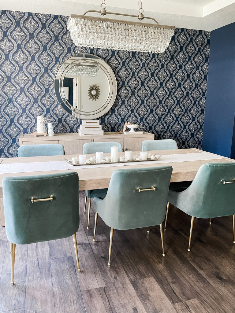 An elegant formal dining space features a long light wood table surrounded by mint velvet dining chairs with gold legs and backplate details. A tray of white candles and flowers runs down the center of the table, under a glamorous rectangular crystal chandelier. The navy blue damask wallpaper continues as a bold backdrop to the mirrored sideboard wall, unifying the design. With a modern glam aesthetic, this Minneapolis-St. Paul design project showcases the boutique studio’s talent for balancing drama with serenity.