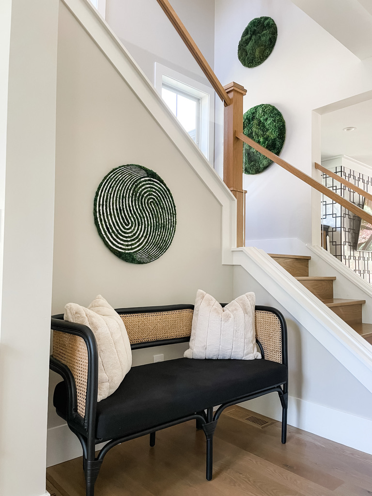 A stairway nook in a Twin Cities home is designed with natural textures and a biophilic style. A black bench with cane siding and plush, cream-colored pillows rests against a soft, white wall. Three green moss wall circles are mounted in an ascending arrangement alongside a wooden handrail. Natural light from small windows enhances the serene, modern-organic ambiance.