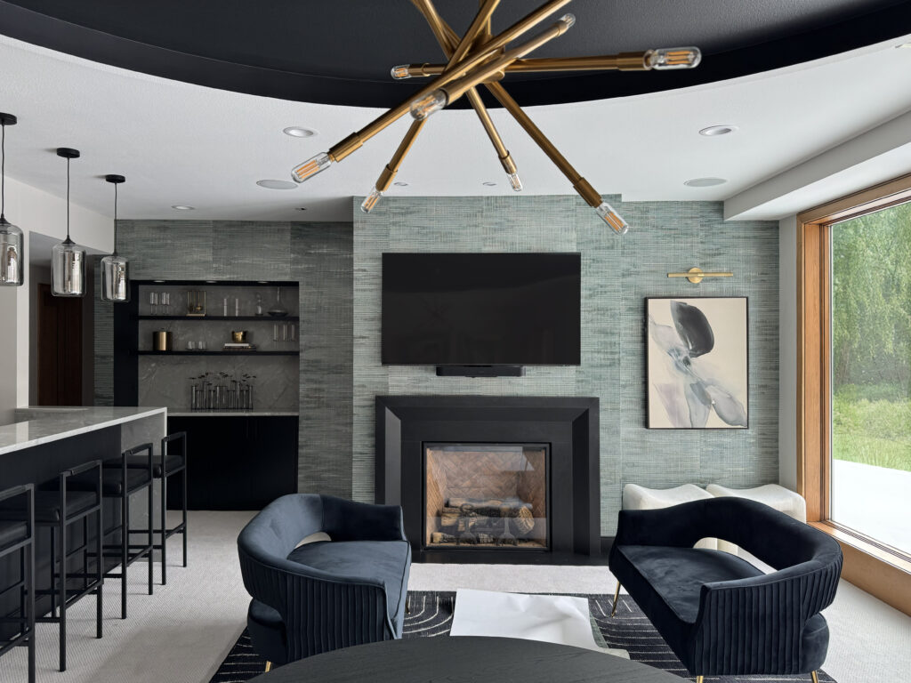A beautiful lower-level entertainment room reimagined. To the left is a white bar with black stools, and three silver light fixtures above it. The main center of the image is a textured gray wallpaper, from the left is a built-in bookshelf (with miscellaneous decorative items on it), in the middle is a TV mounted to the wall above a gas fireplace, a piece of abstract art (with a picture light above it) that's gray with black and compliments the wall. On the far right of the image is a large window that shows a green lush exterior. Int he foreground, a table, and two love seats facing each other with a small table between them.