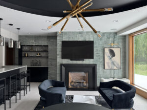 A beautiful lower-level entertainment room reimagined. To the left is a white bar with black stools, and three silver light fixtures above it. The main center of the image is a textured gray wallpaper, from the left is a built-in bookshelf (with miscellaneous decorative items on it), in the middle is a TV mounted to the wall above a gas fireplace, a piece of abstract art (with a picture light above it) that's gray with black and compliments the wall. On the far right of the image is a large window that shows a green lush exterior. Int he foreground, a table, and two love seats facing each other with a small table between them.