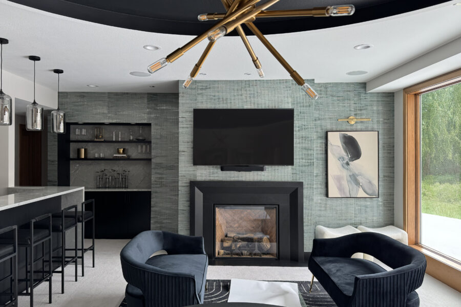 A beautiful lower-level entertainment room reimagined. To the left is a white bar with black stools, and three silver light fixtures above it. The main center of the image is a textured gray wallpaper, from the left is a built-in bookshelf (with miscellaneous decorative items on it), in the middle is a TV mounted to the wall above a gas fireplace, a piece of abstract art (with a picture light above it) that's gray with black and compliments the wall. On the far right of the image is a large window that shows a green lush exterior. Int he foreground, a table, and two love seats facing each other with a small table between them.