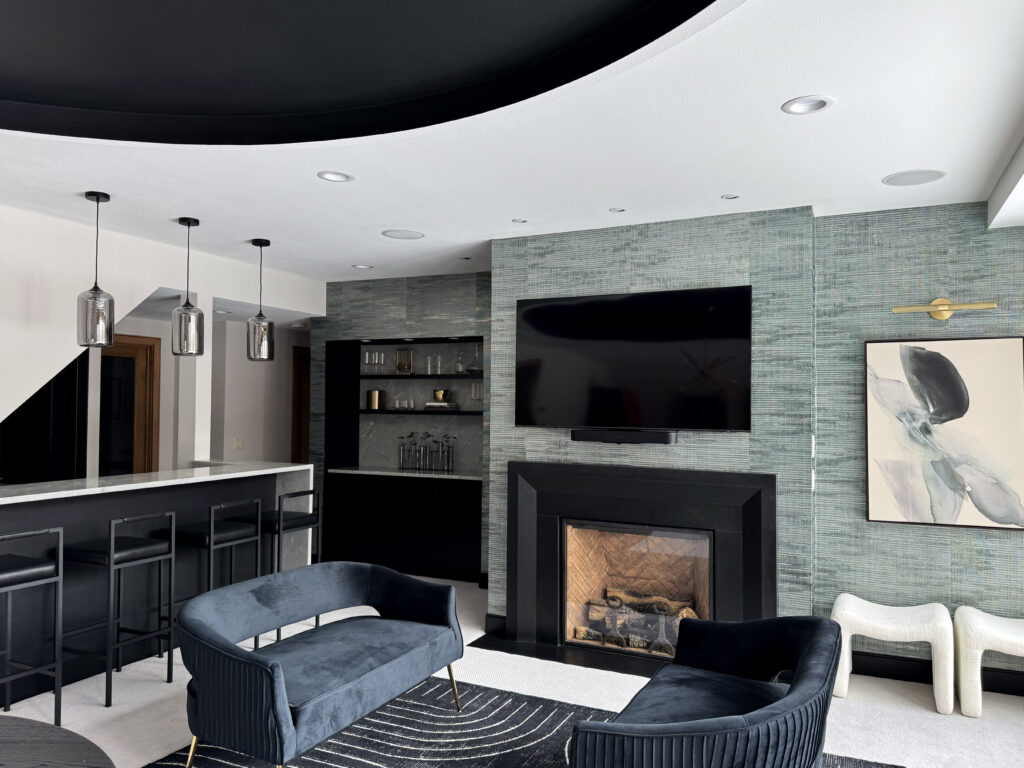 A beautiful lower-level entertainment room reimagined. To the left is a white bar with black stools, and three silver light fixtures above it. The main center of the image is a textured gray wallpaper, from the left is a built-in bookshelf (with miscellaneous decorative items on it), in the middle is a TV mounted to the wall above a gas fireplace, a piece of abstract art (with a picture light above it) that's gray with black and compliments the wall. On the far right of the image is a large window that shows a green lush exterior. Int he foreground, a table, and two love seats facing each other with a small table between them.