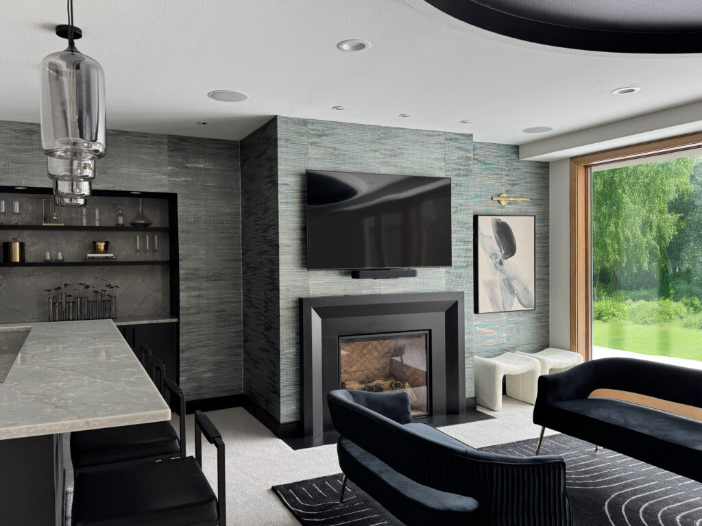 A beautiful lower-level entertainment room reimagined. To the left is a white bar with black stools, and three silver light fixtures above it. The main center of the image is a textured gray wallpaper, from the left is a built-in bookshelf (with miscellaneous decorative items on it), in the middle is a TV mounted to the wall above a gas fireplace, a piece of abstract art (with a picture light above it) that's gray with black and compliments the wall. On the far right of the image is a large window that shows a green lush exterior. Int he foreground, a table, and two love seats facing each other with a small table between them.