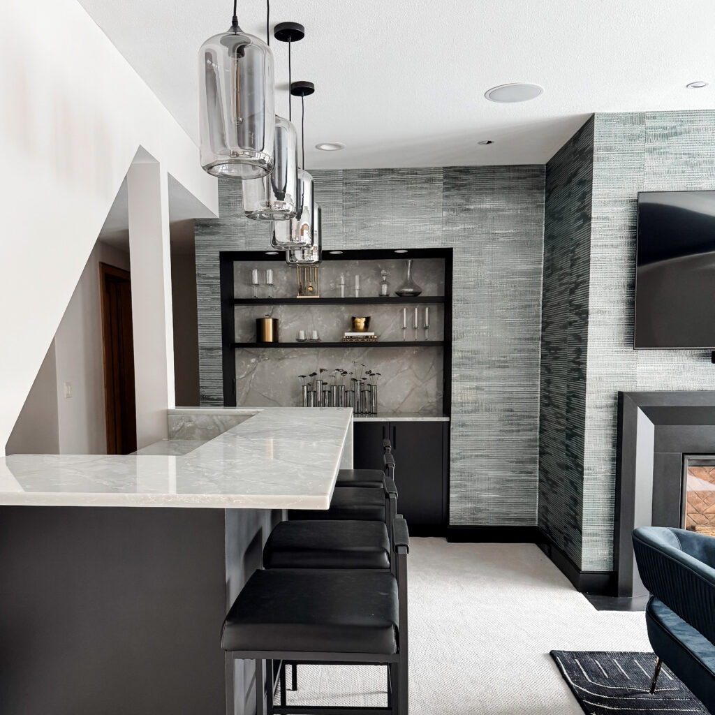 A beautiful lower-level entertainment room reimagined. To the left is a white bar with black stools, and four silver light fixtures above it. Off center of the image, but the background, is a textured gray wallpaper, from the left is a built-in bookshelf (with miscellaneous decorative items on it), on the right is a TV mounted to the wall above a gas fireplace. In the foreground, a is a blue love seat.
