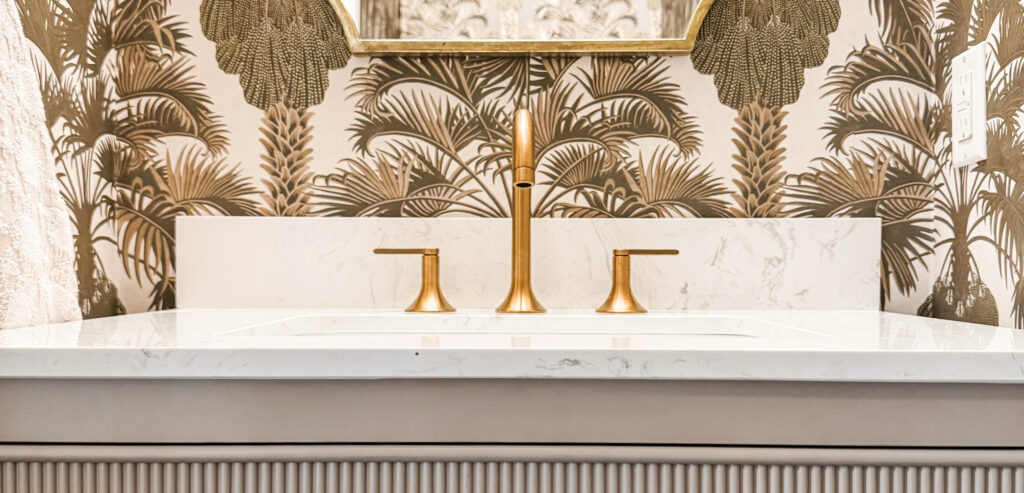 Closeup of a sink with a brass faucet, a white marble countertop, and a gray exterior. Wallpaper in the background with palm trees.