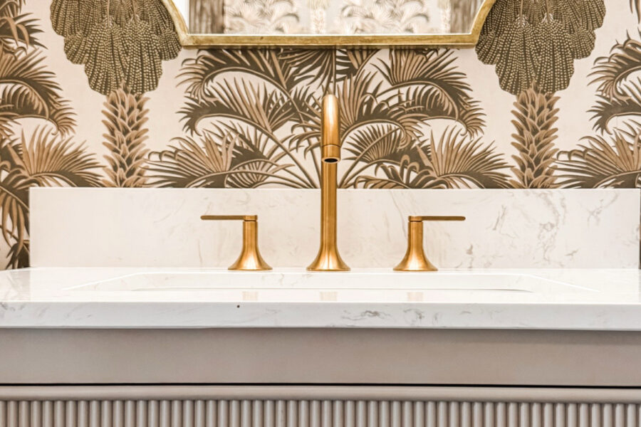 Closeup of a sink with a brass faucet, a white marble countertop, and a gray exterior. Wallpaper in the background with palm trees.