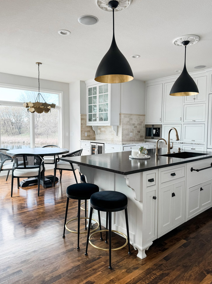 Bright, modern kitchen in the Minneapolis-St. Paul area featuring white custom cabinetry, a dark quartz island countertop with built-in sink and gold-tone faucet, and matte black pendant lights with gold interiors. The island includes three black-cushioned stools, and the adjacent dining area showcases a round table with black-framed chairs and a sculptural gold chandelier. Large windows provide a serene view of leafless trees and a reflective pond. This kitchen design highlights the creative vision and boutique approach of the interior design team, who blend elegance with function for custom homes throughout the Twin Cities and surrounding suburbs.