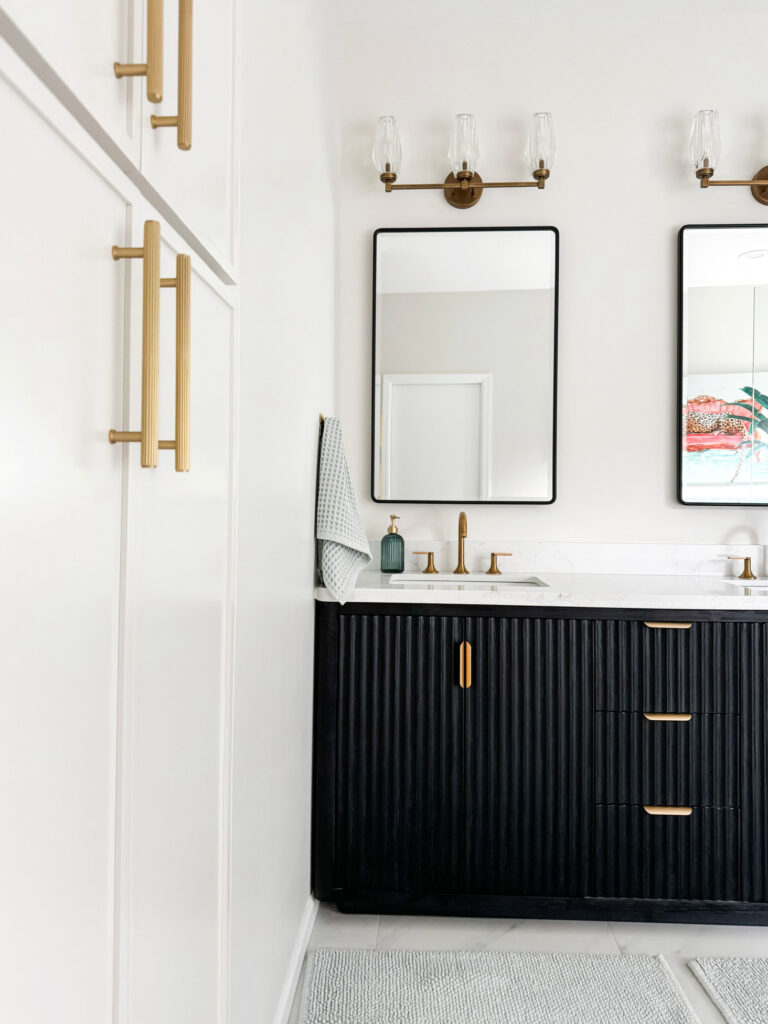 This alternate angle of the Minneapolis primary bathroom shows the elegant white-paneled cabinetry with oversized brushed brass handles. Twin mirrors above the black fluted vanity enhance the symmetry and sophistication, accented by three-globe wall sconces. From fixture finishes to custom cabinetry, this interior design team’s creative approach brings high-end function and flair to Twin Cities bathrooms.