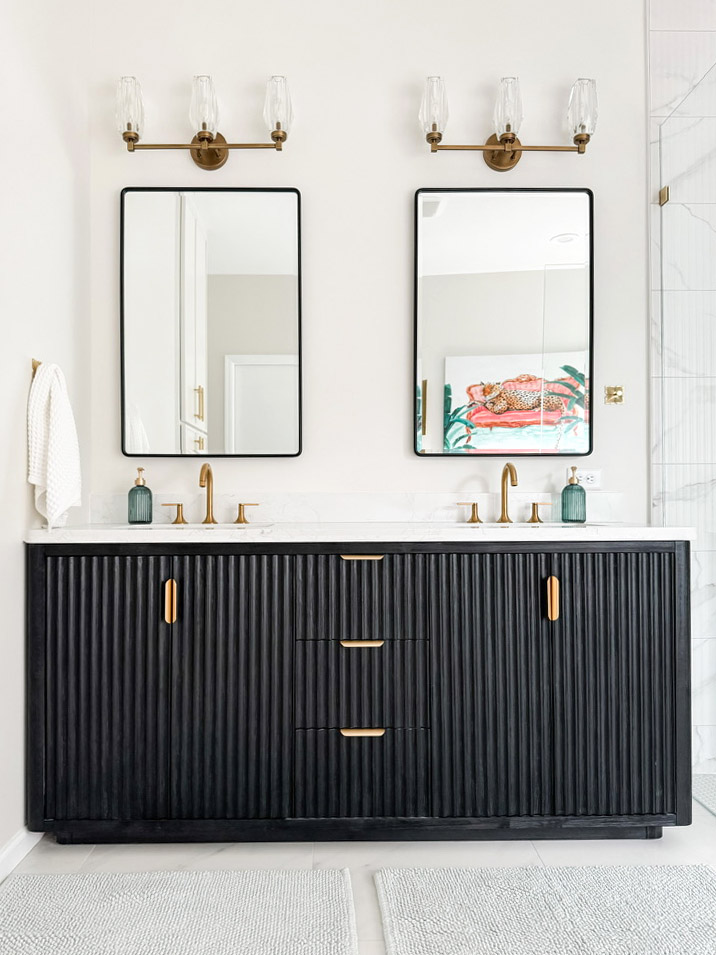 Firelight Style House redesigned the primary bathroom in Wayzata. The image is of the dual sink vanity, with a brass faucet and handles. The vanity is black and textured with brass handles. There are mirrors above the two sinks, and lights mounted above the mirrors with three lights each. Wonderful lighting design for this primary bathroom remodel.