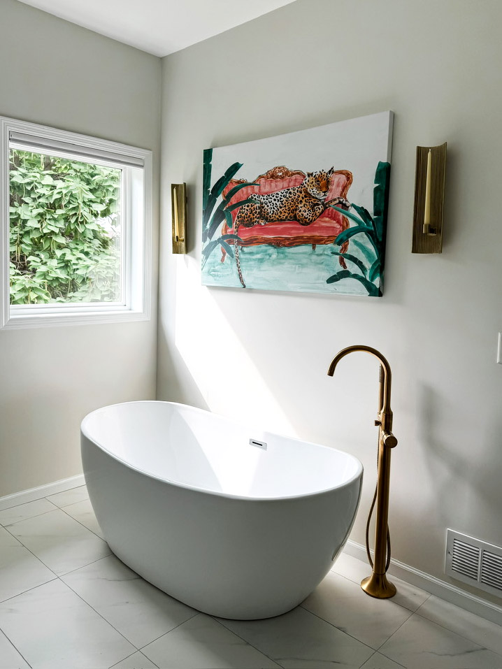 Firelight Style House redesigned the primary bathroom in Wayzata. The image shows a beautiful large bathtub that sits alone and away from the wall. Next to it is the faucet, which also stands alone and is in brass. Above the tub is a painting of a leopard lying on a red couch. On both sides of the art are wall sconces for candles. There's a square window on the left side of the tub.