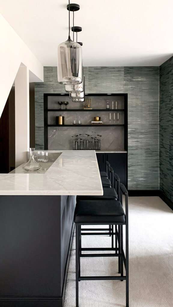 A beautiful lower-level entertainment room reimagined. To the left is a white bar with black stools, and four silver light fixtures above it. Off center of the image, but the background, is a textured gray wallpaper.