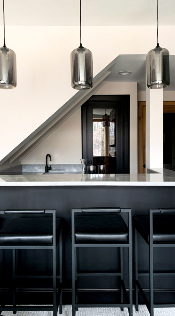 Modern black kitchen bar with three leather barstools, a marble countertop, matte black cabinetry, and smoky glass pendant lights beneath a sloped ceiling.