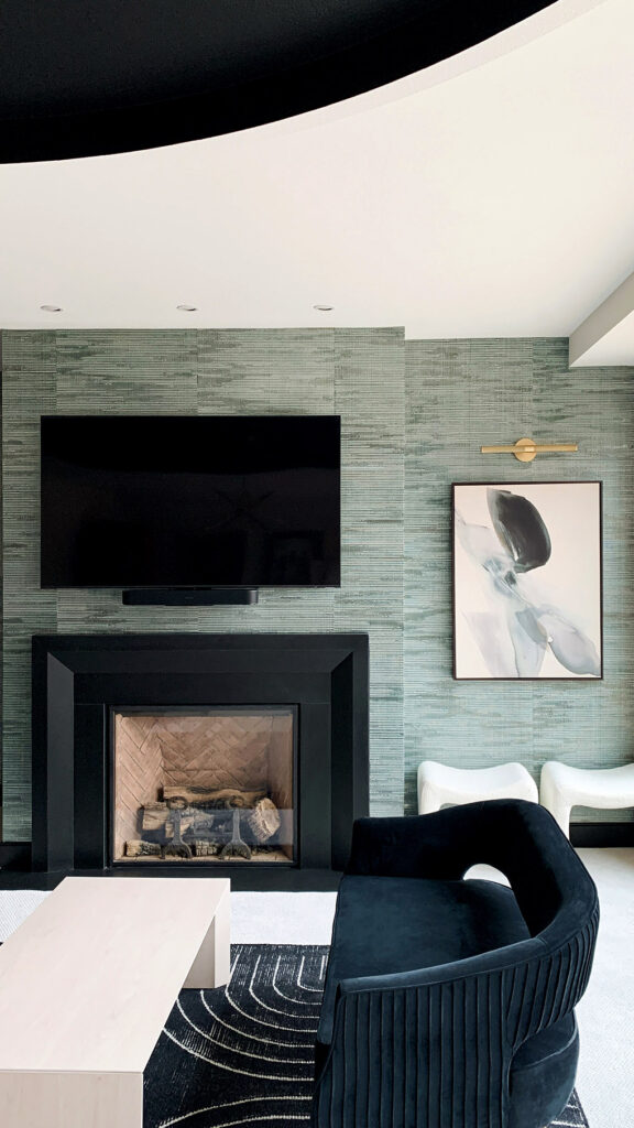 A beautiful lower-level entertainment room reimagined. Off center of the image, but the background, is a textured gray wallpaper, centered is a a TV mounted on the wall above a gas fireplace. In the foreground, a is a blue love seat with white table on a blue rug with small white stripes..
