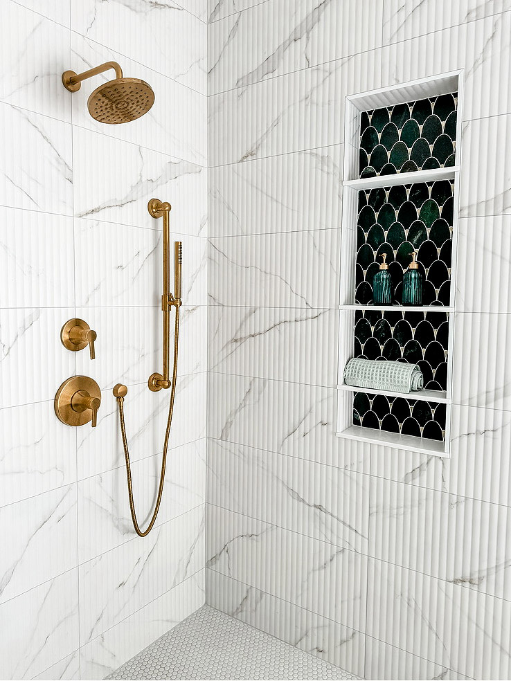 Firelight Style House redesigned the primary bathroom in Wayzata. The image shows a large primary shower with new, textured white marble tiles. There is a large copper shower head, matching levers, and an extendable mini shower head. There's a built-in cubby area to hold shampoo, featuring an arched black tile in the back wall. It has four shelves, with one holding a rolled-up towel and the other containing two small glass pump bottles.