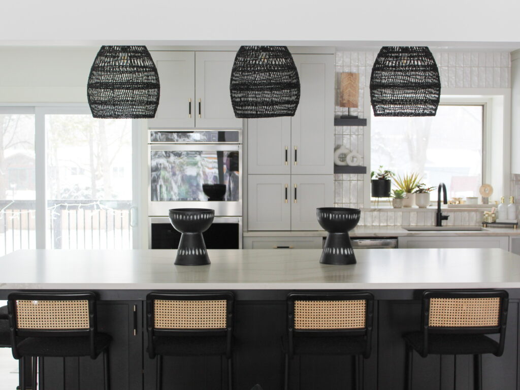 A newly remodeled Minneapolis-St. Paul area kitchen features black woven pendant lights, a large island with cane-back stools, and sleek white cabinetry with gold hardware. There's a window behind the sink and a sliding door to a patio on the right.