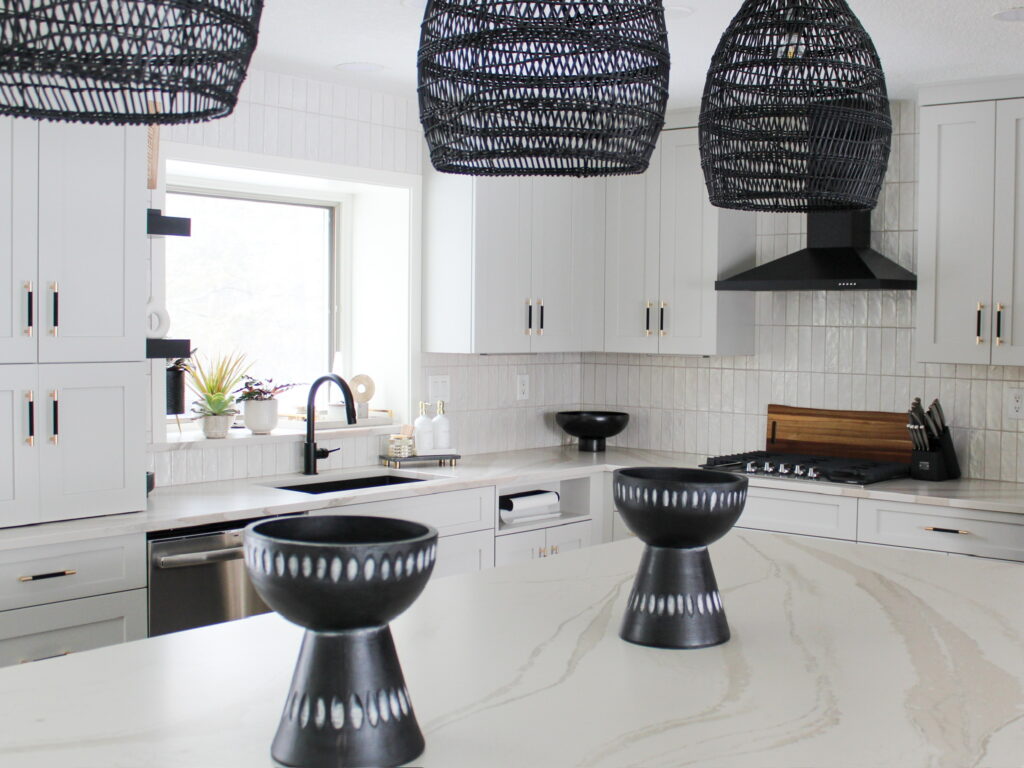 Bright and airy kitchen remodel in a Minneapolis suburb showcasing creamy white cabinetry, a sculptural matte black hood, and rattan pendant lighting above a waterfall-edge quartz island. The Firelight Design team blended bold textures and neutral tones with custom lighting and storage solutions that reflect their boutique, client-focused approach.