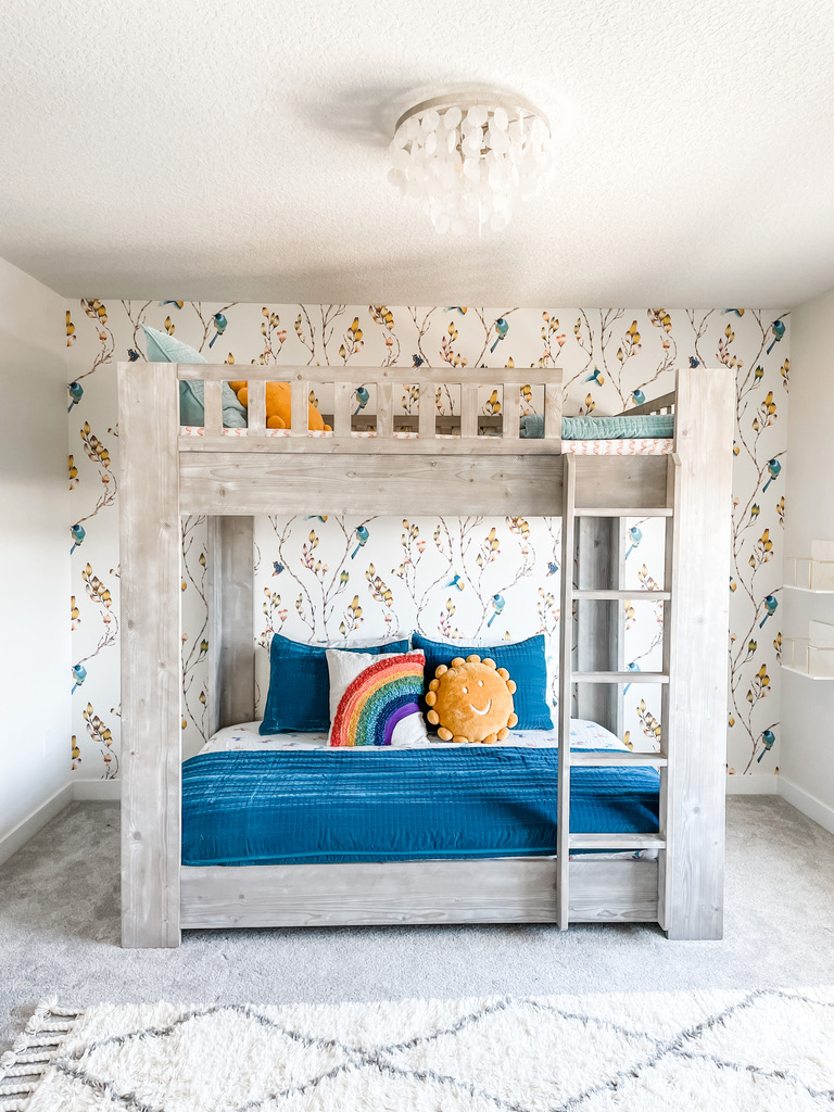 This charming children’s room in a Twin Cities home highlights a wood-finish bunk bed with plush teal bedding, set against a whimsical wallpaper featuring delicate yellow buds and perched bluebirds. A large cream and gray diamond-pattern rug softens the floor, while a dresser and woven lamp add texture and style.