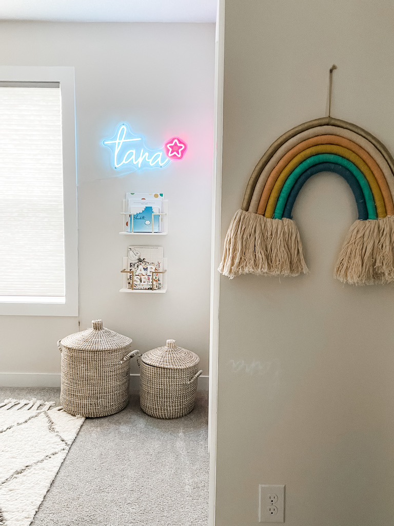 Kids room corner with a woven rainbow wall hanging, two lidded wicker baskets on the floor, a small book display, and a glowing neon name sign near a window.