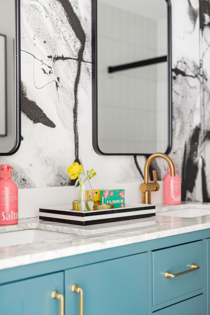 Detail of Minneapolis-St. Paul's double vanity features a gold faucet, teal cabinetry, and a striped tray styled with bright accents and fresh florals.