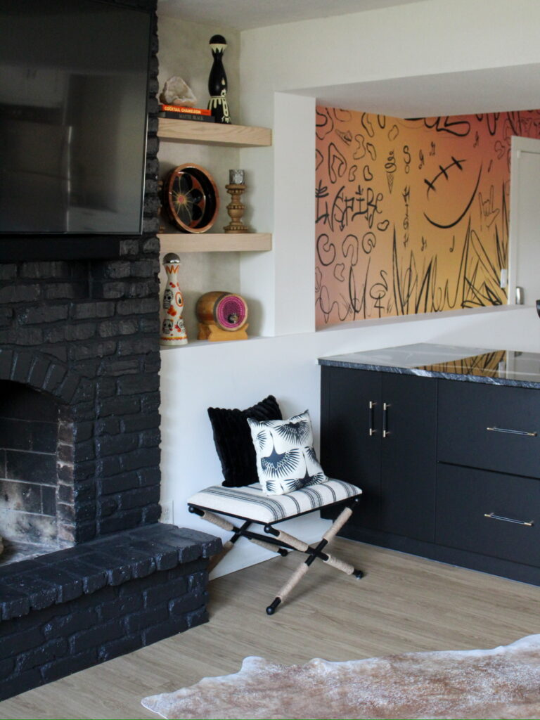 An eclectic living space revisioned in the Twin Cities area features a matte black, painted brick fireplace, open wood shelves with global decor, and a warm, abstract mural, all contributing to a vibrant character.