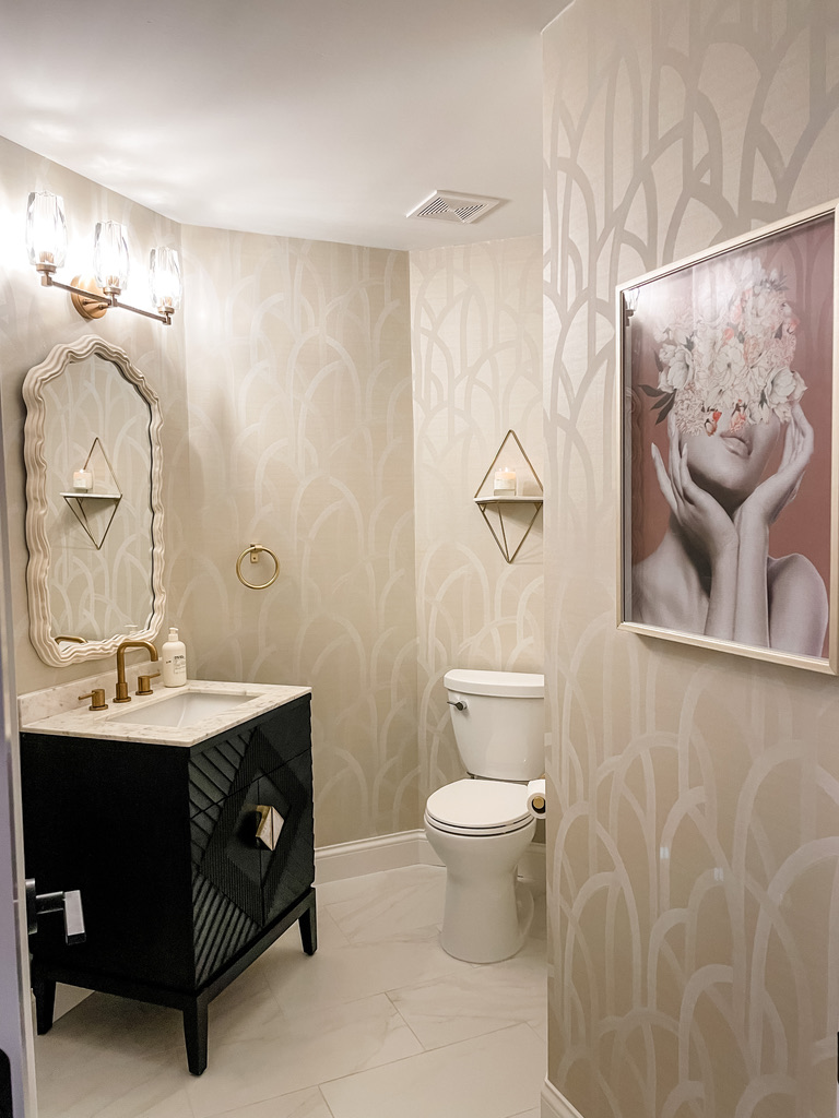 Minneapolis-St. Paul's powder room has soft metallic wallpaper, a scalloped mirror, a black vanity, and gold fixtures for a glam touch.