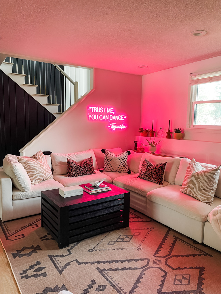 A fun and inviting lower level in a remodeled home in Minneapolis features a cozy white sectional, bold accent pillows, and a hot pink neon sign reading “Trust Me, You Can Dance – Tequila,” radiating playful energy and a warm, inviting feel, courtesy of the Firelight interior design team.