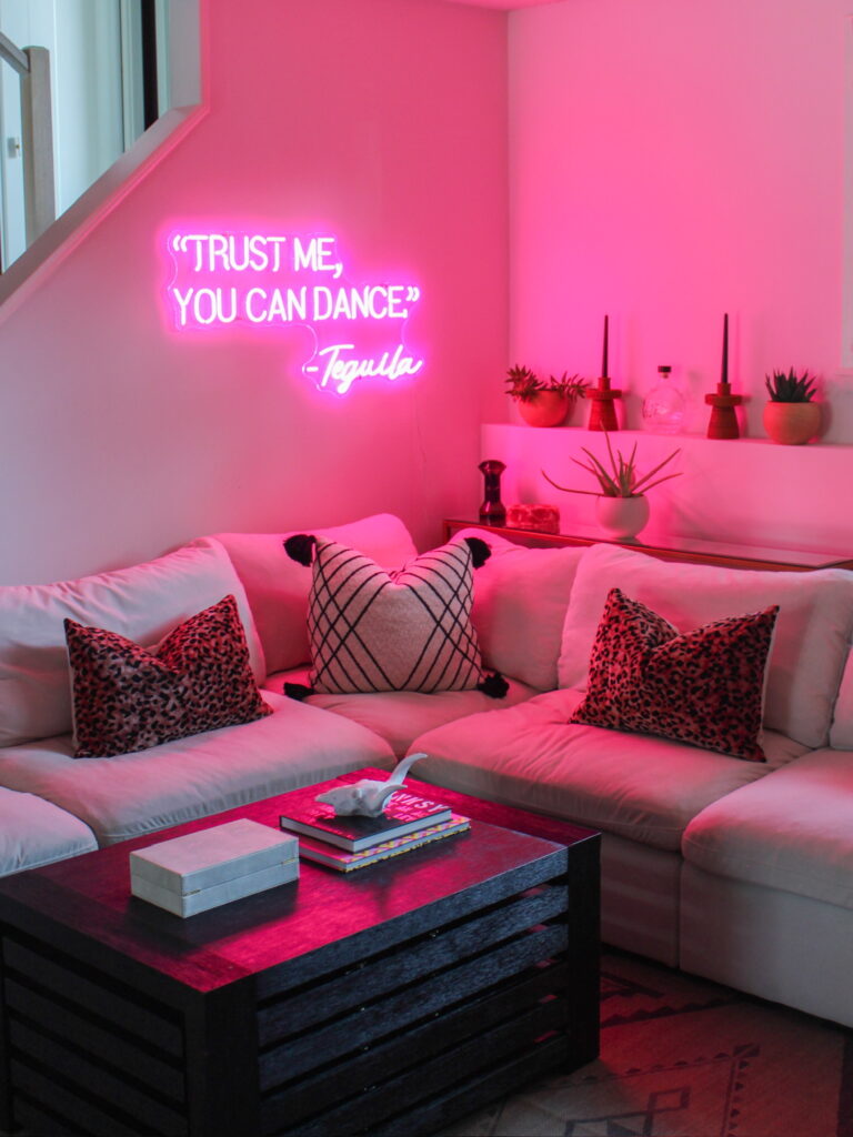 A cozy corner in a Minneapolis-St. Paul home glows with pink neon reading “Trust Me, You Can Dance – Tequila,” above a white sectional with leopard and black-trimmed pillows, potted succulents, and a dark wood coffee table—a fun interior design aesthetic completed by the Firelight Design Team.