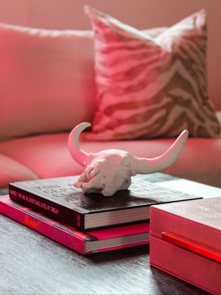 A horned white ceramic skull sits atop design books on a black wood table, lit by soft pink light in this Minneapolis-St. Paul lower level home that Firelight Style House remodeled. A zebra-patterned pillow adds playful texture against the creamy sectional.