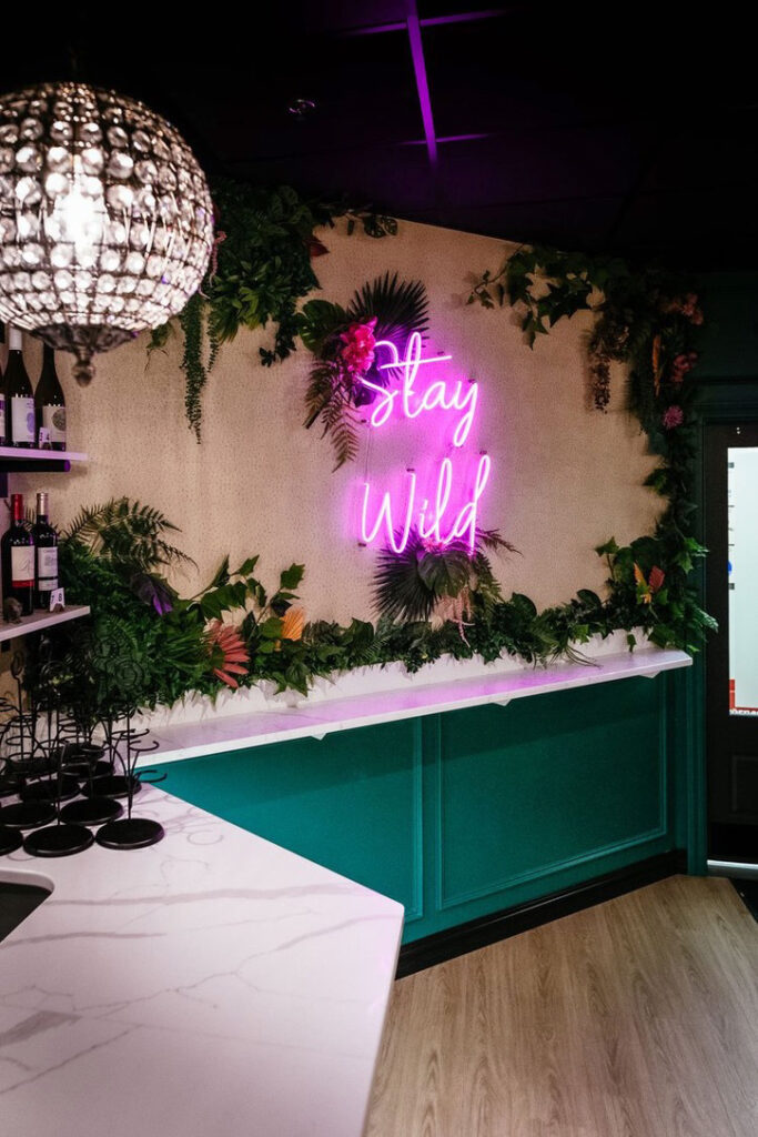 A vibrant corner of Rhino Wine Bar in the Minneapolis-St. Paul area featuring a lush green wall of tropical plants and a neon pink sign that reads “Stay Wild.” The bar’s white marble counter contrasts beautifully with emerald green paneling and a sparkling crystal pendant light, creating an inviting and bold atmosphere.