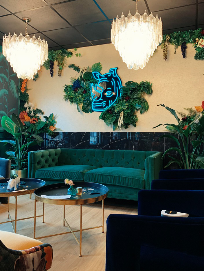 A chic lounge area in Rhino Wine Bar, located near Minneapolis, showcasing a deep emerald green tufted velvet couch set against a lush living wall and a glowing blue neon rhino head. The room is styled with bold greenery, glamorous chandeliers, and modern gold-accented tables.