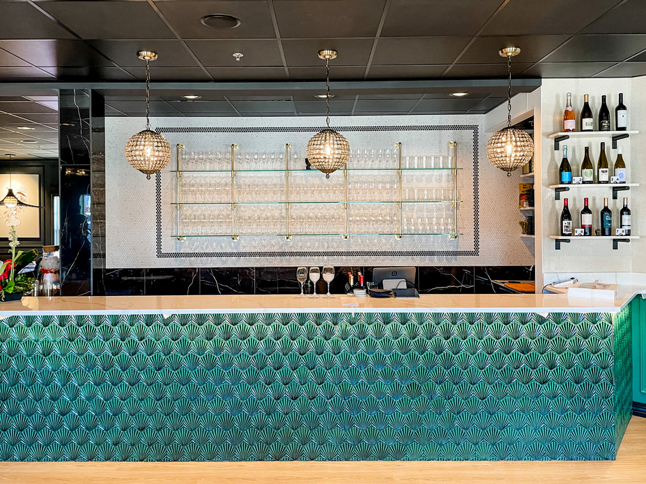 A vibrant wine bar counter in the Minneapolis-St. Paul area features a bold emerald green tile front with an art deco fan pattern, topped by a sleek white stone countertop. Above the bar, three crystal globe pendant lights hang in a row, illuminating a mirrored backdrop with brass-accented glass shelving filled with wine glasses. Bottles of wine are neatly arranged on floating shelves to the right, adding warmth and charm to the elegant setting.