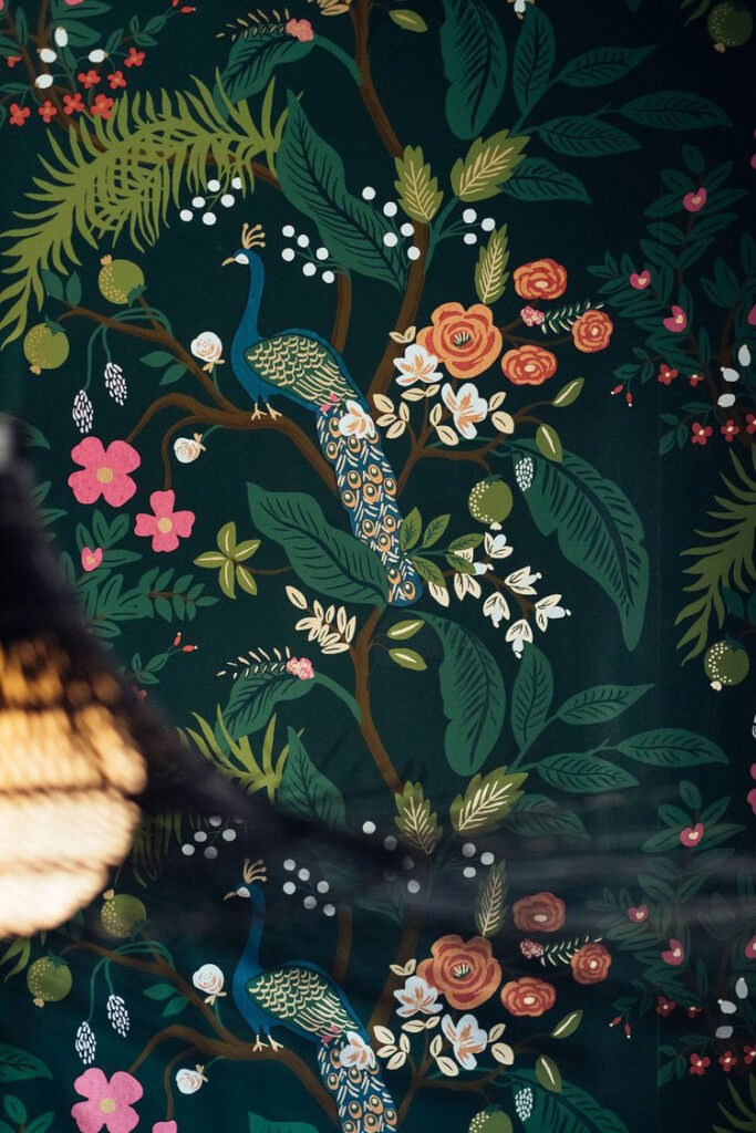 A close-up of botanical wallpaper in Rhino Wine Bar shows an illustrated peacock perched among blooming flowers and lush green leaves. The peacock’s teal and gold plumage contrasts beautifully with the deep forest green background, creating a rich, inviting atmosphere reflective of Minneapolis-St. Paul’s thriving design scene. The interior design team’s boutique approach shines in this custom-selected wall covering, thoughtfully chosen to elevate the bar's distinctive, whimsical style.