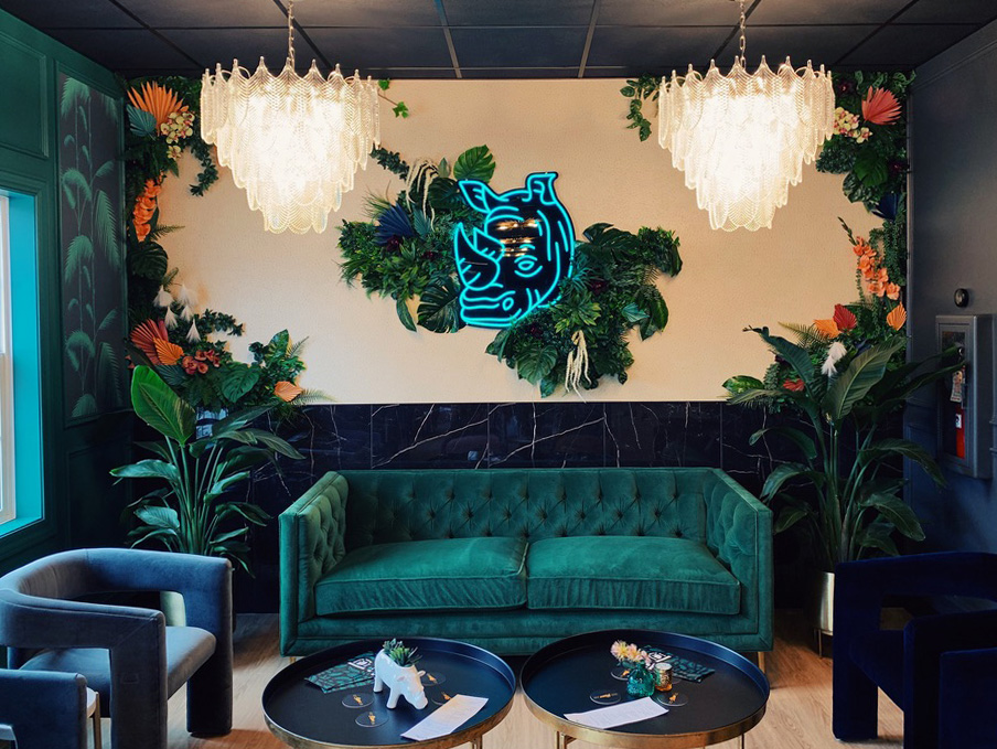 A wide view of the Rhino Wine Bar’s signature lounge area, featuring a bold green velvet sofa flanked by leafy tropical plants and twin crystal chandeliers. The neon blue rhino logo anchors a lush, greenery-framed accent wall, blending playful branding with refined interior design.