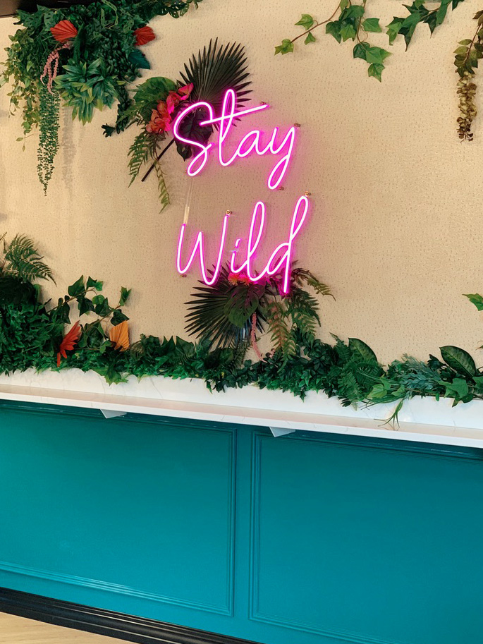 A bold feature wall at Rhino Wine Bar in Minneapolis showcases a hot pink neon 'Stay Wild' sign surrounded by lush tropical greenery. The teal wainscoting and marble countertop add vibrant contrast and modern sophistication to the space.