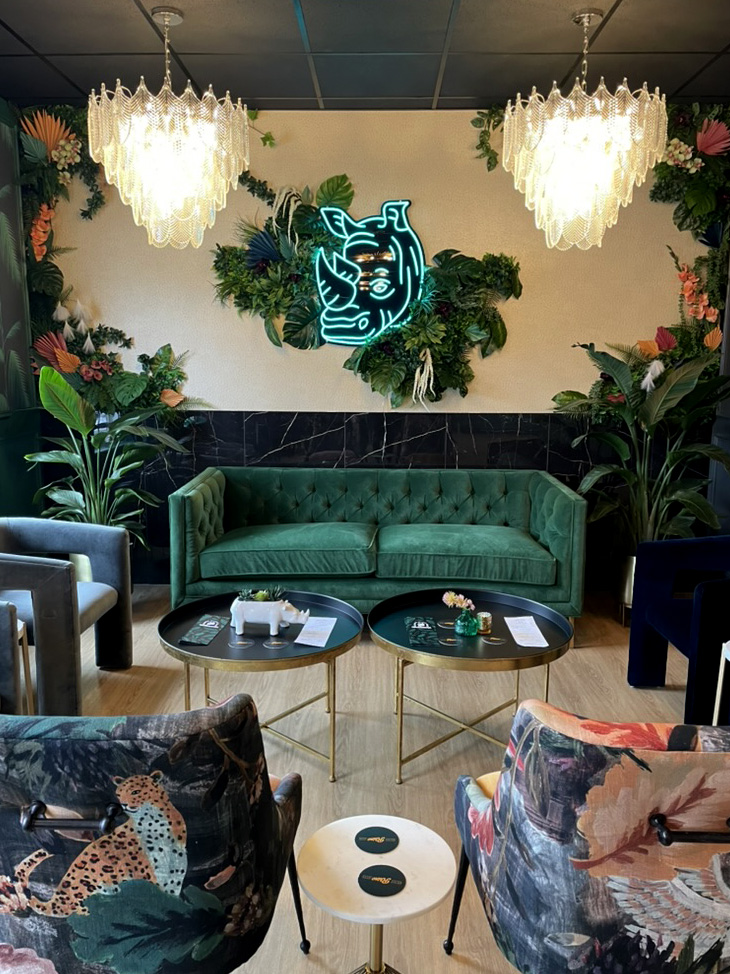 This cozy seating area at Rhino Wine Bar features a plush emerald green velvet couch set against a wall adorned with greenery and a bright blue neon rhino head. Two sculptural crystal chandeliers hang above, reflecting the eclectic elegance of this St. Paul favorite.