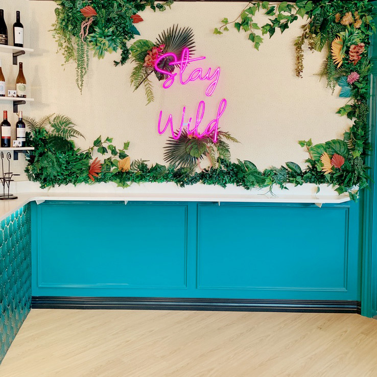 A bold feature wall at Rhino Wine Bar in Minneapolis showcases a hot pink neon 'Stay Wild' sign surrounded by lush tropical greenery. The teal wainscoting and marble countertop add vibrant contrast and modern sophistication to the space.