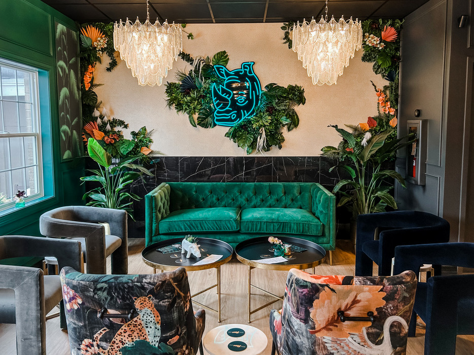 With twin crystal chandeliers and a deep green velvet couch under a tropical plant wall, this Rhino Wine Bar lounge exudes luxe energy. A glowing blue neon rhino is the design centerpiece, framed by a canopy of foliage that feels both wild and curated.