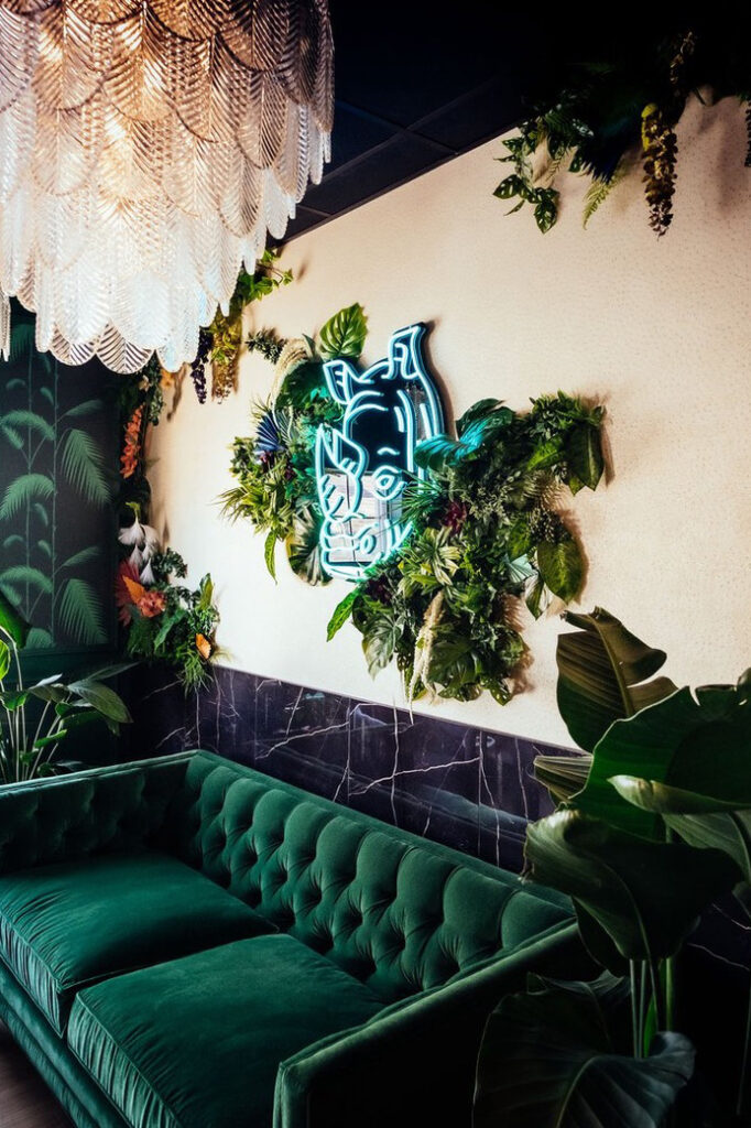 An angled view of Rhino Wine Bar’s signature rhino neon installation highlights the texture of greenery framing the electric blue art. This bold statement wall complements the tufted velvet seating below, demonstrating the design team's custom flair and fearless use of texture and light.
