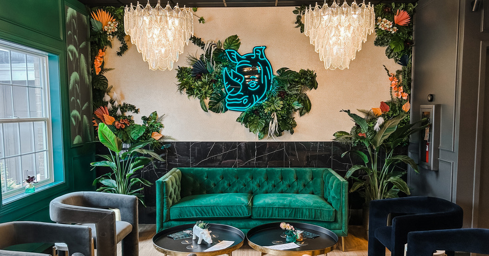 A modern, tropical-themed lounge interior designed by the Firelight design team, featuring a luxurious green velvet couch, elegant chandeliers, and lush plant décor. Vibrant floral arrangements frame a glowing neon rhinoceros sign, creating a bold yet inviting vibe. Velvet armchairs, black marble accents, and natural light complete the sophisticated atmosphere—perfectly blending interior design, comfort, and visual storytelling.
