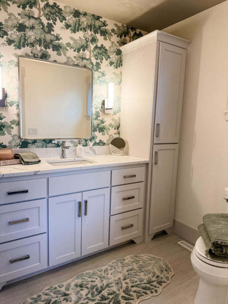 Bright bathroom with a white vanity and quartz countertop, tall linen cabinet, and rectangular mirror, accented by green-and-cream botanical wallpaper, modern wall sconces, light wood-look tile flooring, and a soft green patterned bath rug.