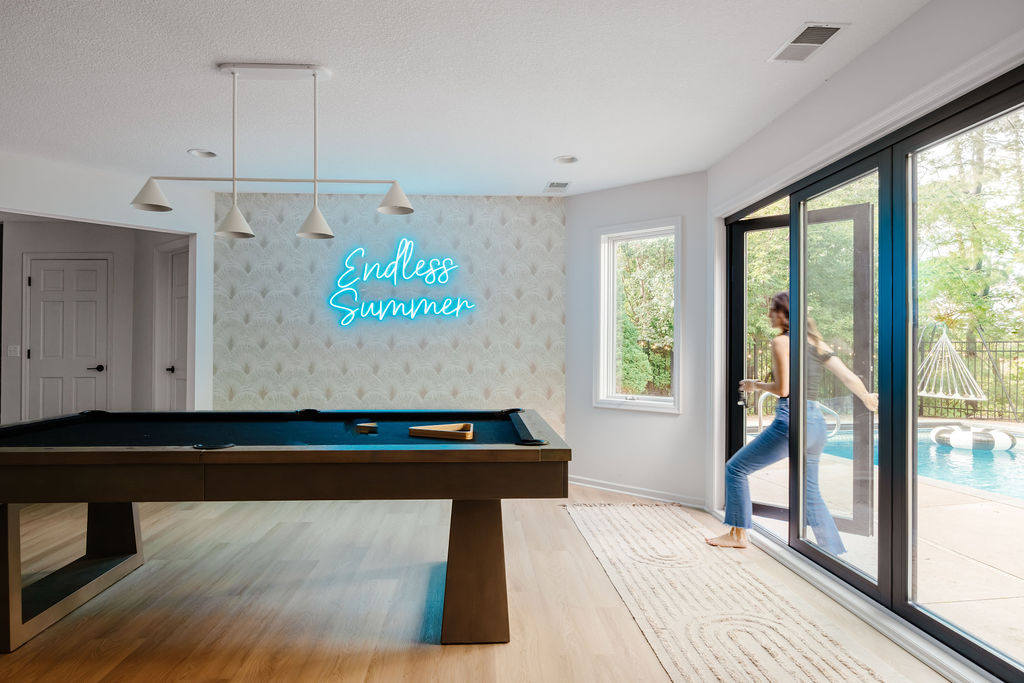 A women entering a bright, modern game room with a black pool table, light wood floors, white pendant lights, and patterned wallpaper featuring a neon-style sign that reads “Endless Summer.”