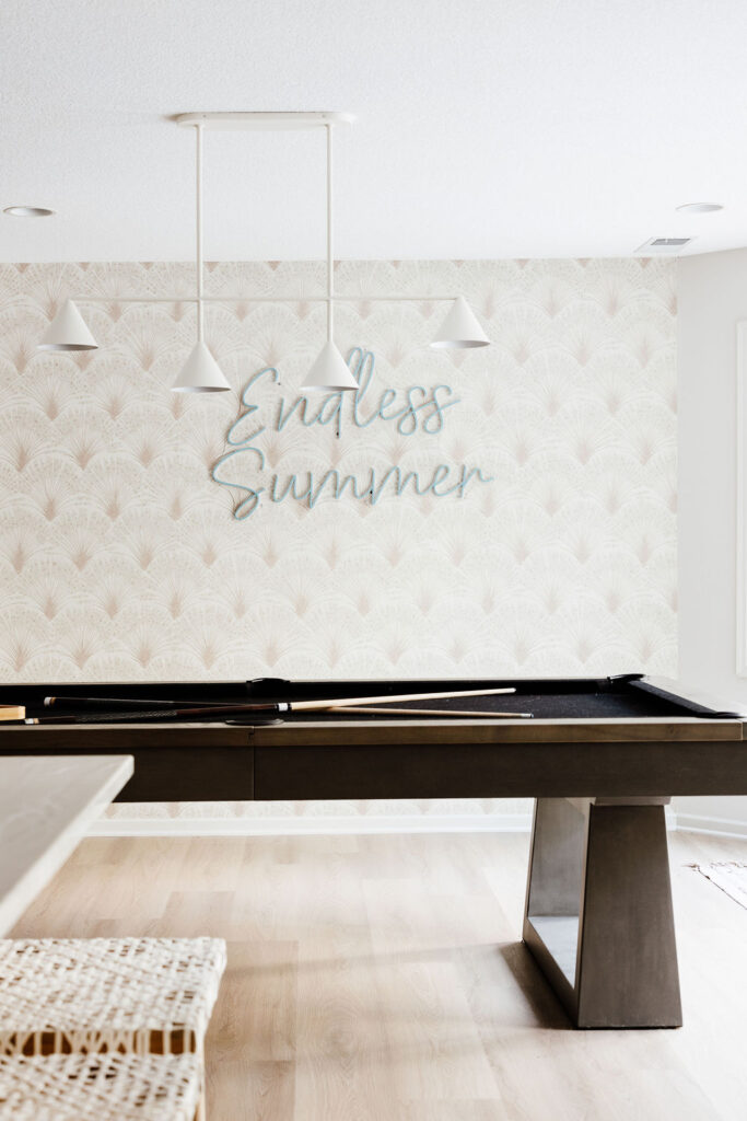 Bright, modern game room with a black pool table, light wood floors, white pendant lights, and patterned wallpaper featuring a neon-style sign that reads “Endless Summer.”