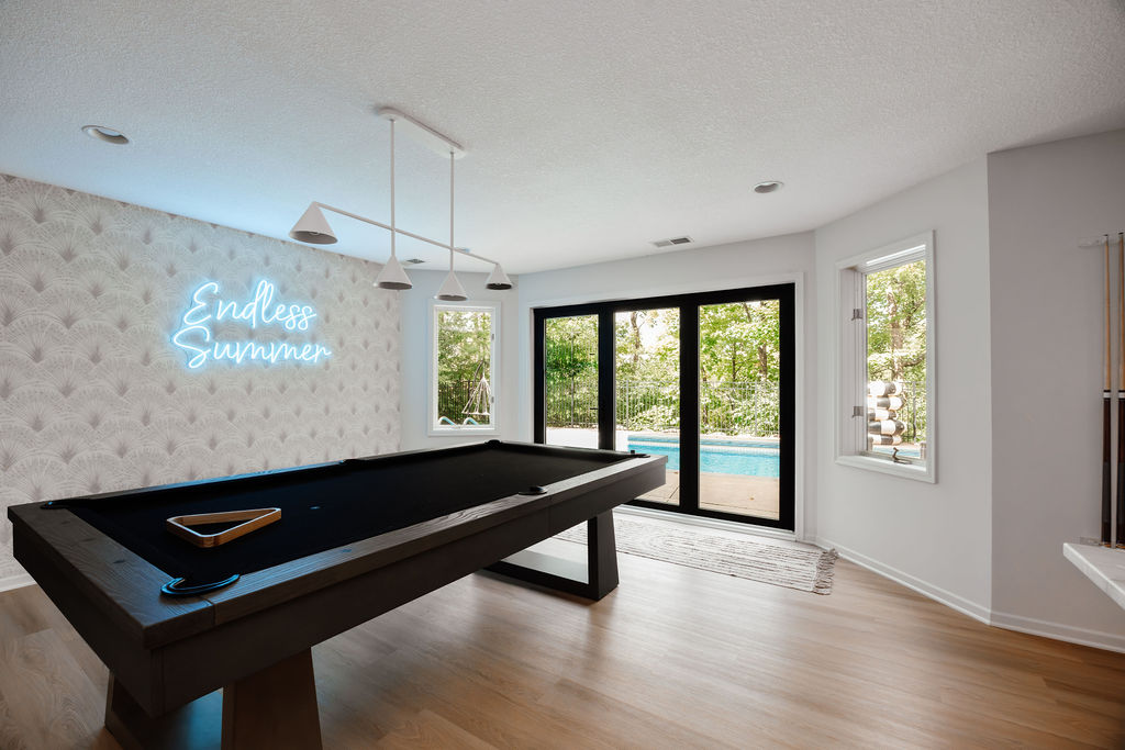 Bright, modern game room with a black pool table, light wood floors, white pendant lights, and patterned wallpaper featuring a neon-style sign that reads “Endless Summer from a different angle.